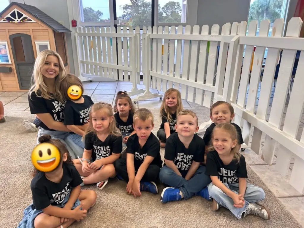 Preschool students at Bethany Christian Preschool smiling with their teacher, wearing matching "Jesus Loves You" shirts inside a bright classroom.