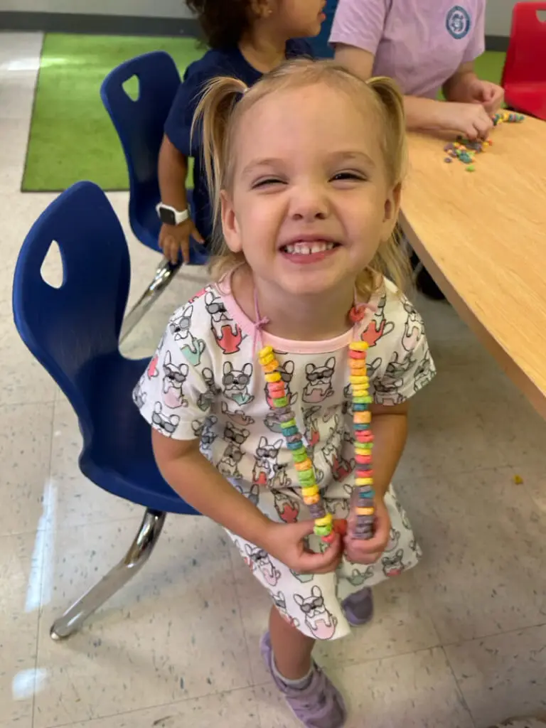 Smiling preschool girl holding a colorful cereal necklace during a classroom activity at Bethany Christian Preschool in West Melbourne, FL.